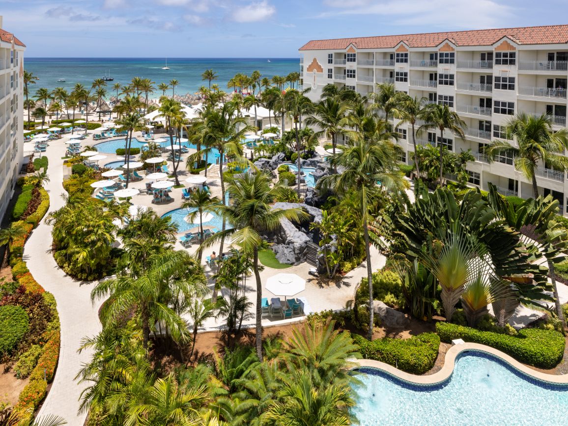 Exterior of Marriott's Aruba Ocean Club timeshare, swimming pool under a canopy of palm trees, ocean in the background